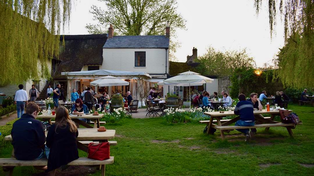 People enjoy drinks in a beautiful pub garden surrounded by weeping willows.