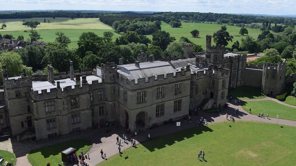 Amidst green fields dotted with trees, Warwick Castle appears on a sunny day, with people exploring the courtyard in front of the Great Hall.