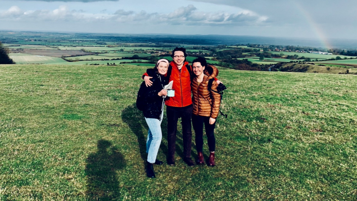Three students stand on top of a hill with a beautiful view of the surrounding countryside behind them.