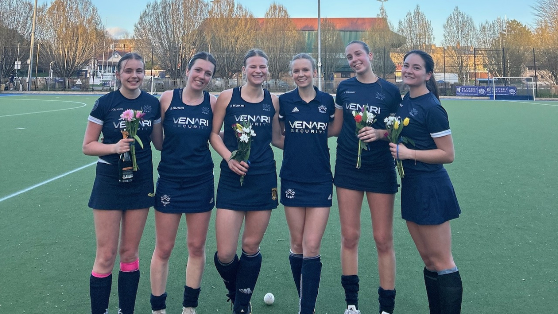 Six students stand smiling on a field hockey pitch in Oxford holding flowers having just won their game.