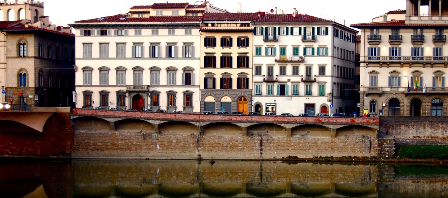 Old European-style apartment buildings line a street overlooking the Arno River in Florence. The buildings' reflection are clearly shown in the river water.