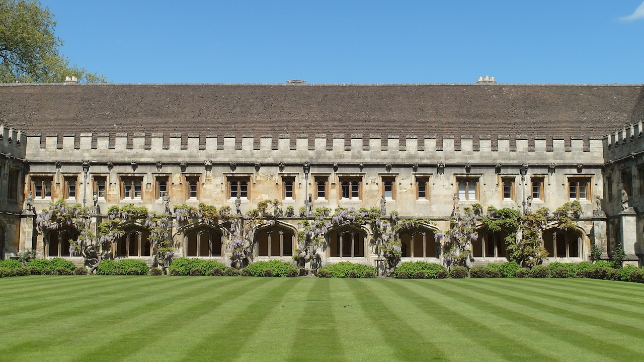 Magdalen College on a sunny Spring day. There is a beautiful blue sky, an immaculately kept lawn and archways festooned with wisteria.