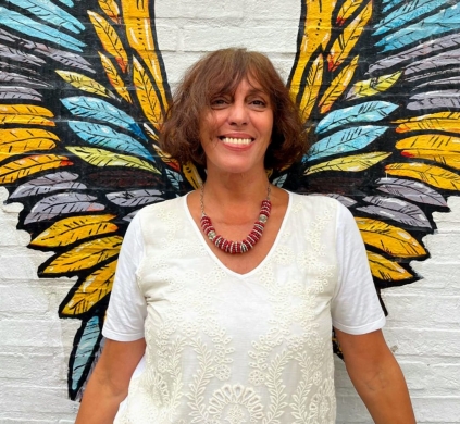 A woman with short, dark hair, a red beaded necklace, and white t-shirt stands in front of a pair of painted wings smiling