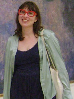A young woman with brown hair and red glasses stands before a painting in a green sweater, black tee-shirt, and black pants