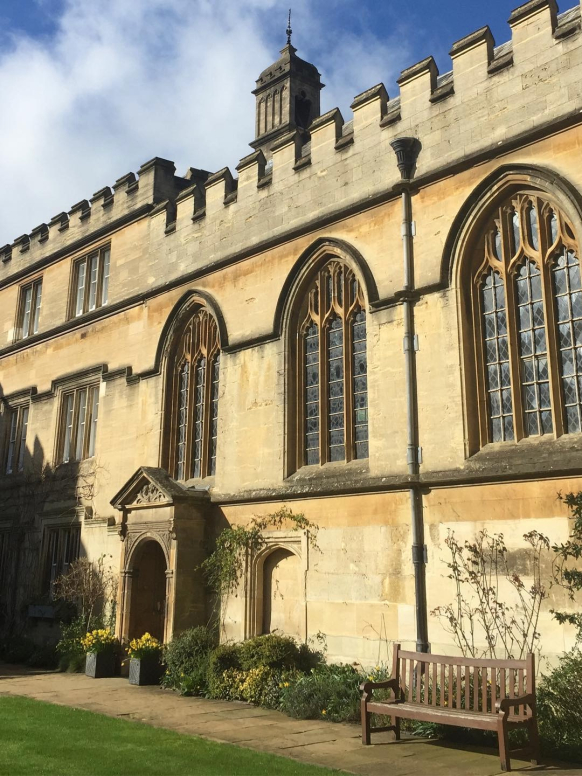 The golden stone of an Oxford College shines with Spring sunshine. Daffodils are seen in planters and a bench tempts people to take a pause and enjoy the warmth.