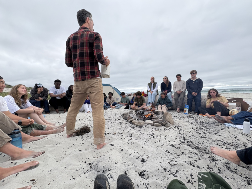 Students sit around a bonfire on a beach while an instructor stands holding a watering can
