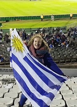 A student stands in a soccer stadium, holding the flag of Uruguay