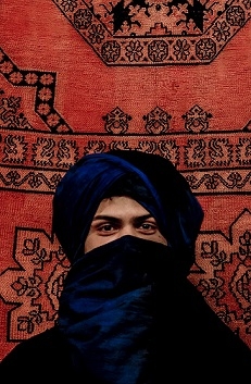 A student with a Berber scarf around his face, in front of a woven rug