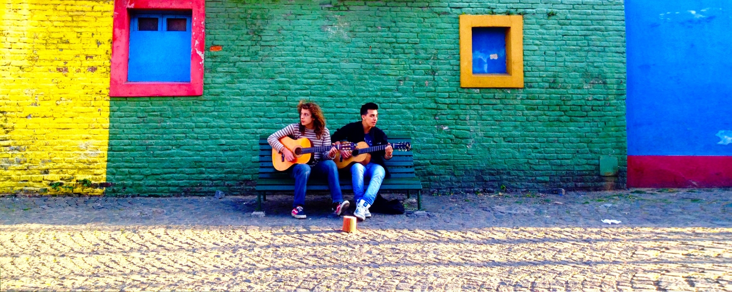 Male and female student sitting in front of colorful wall, playing guitar