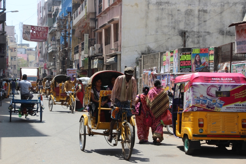A man transports women in a rickshaw while others drive and walk by