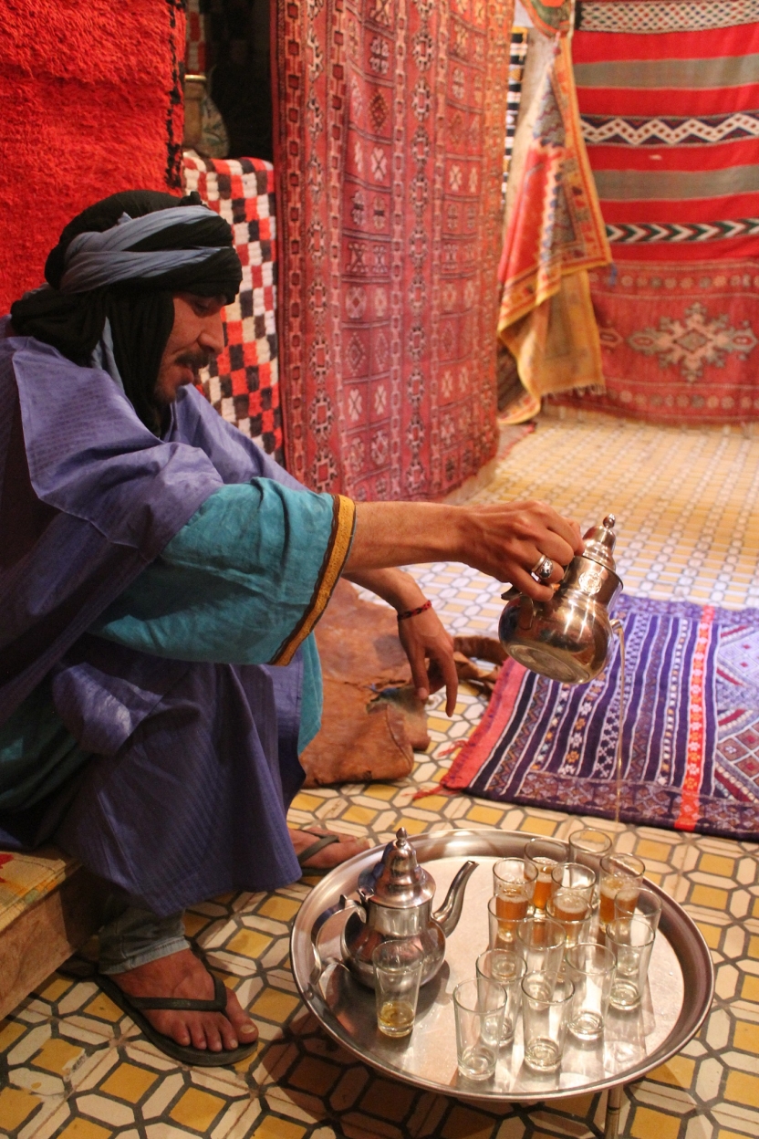 A man pours tea with rugs behind him