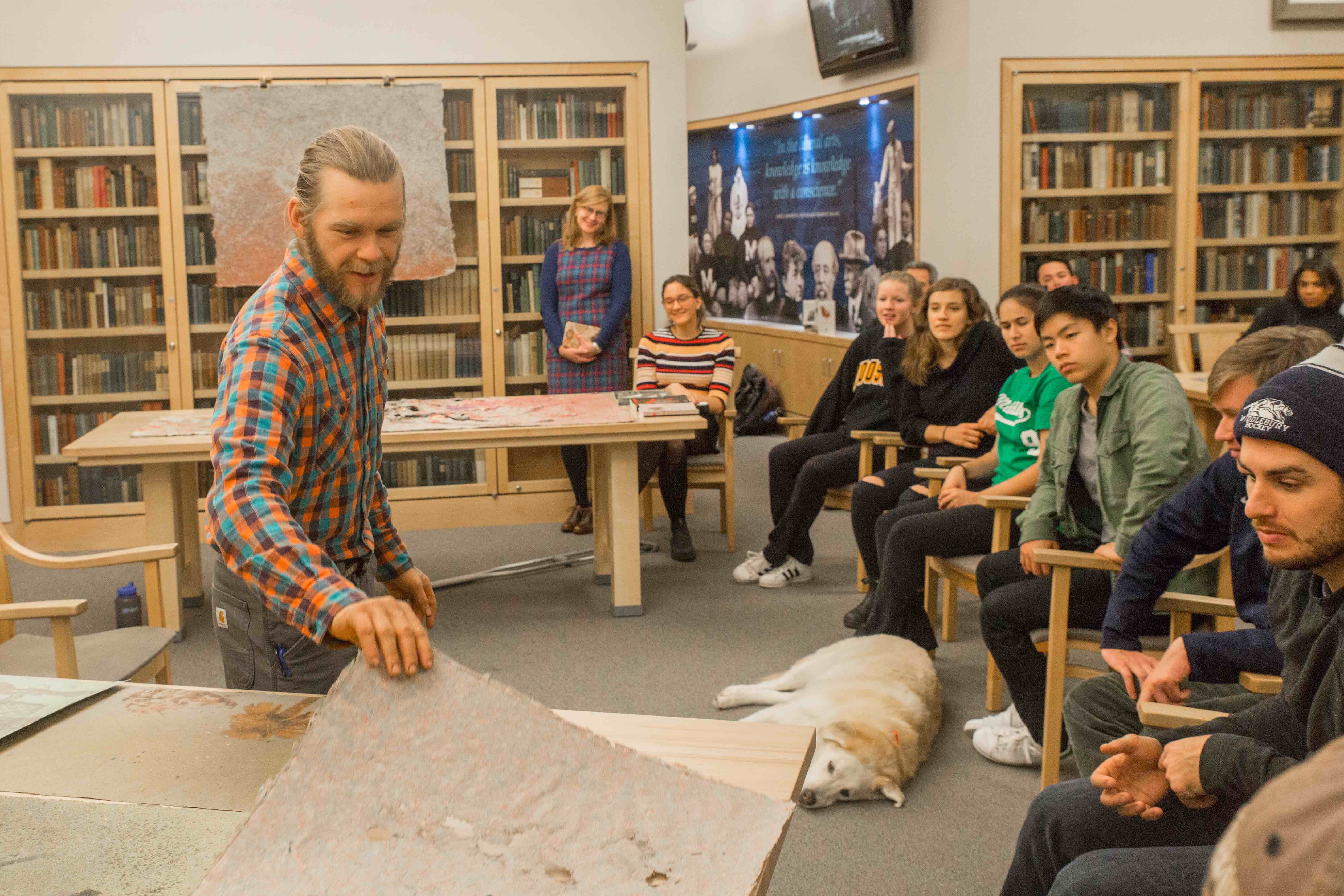 Students look on attenvtiely as the lecturer lifts up a piece of art from a table. A dog also lies on the ground. 