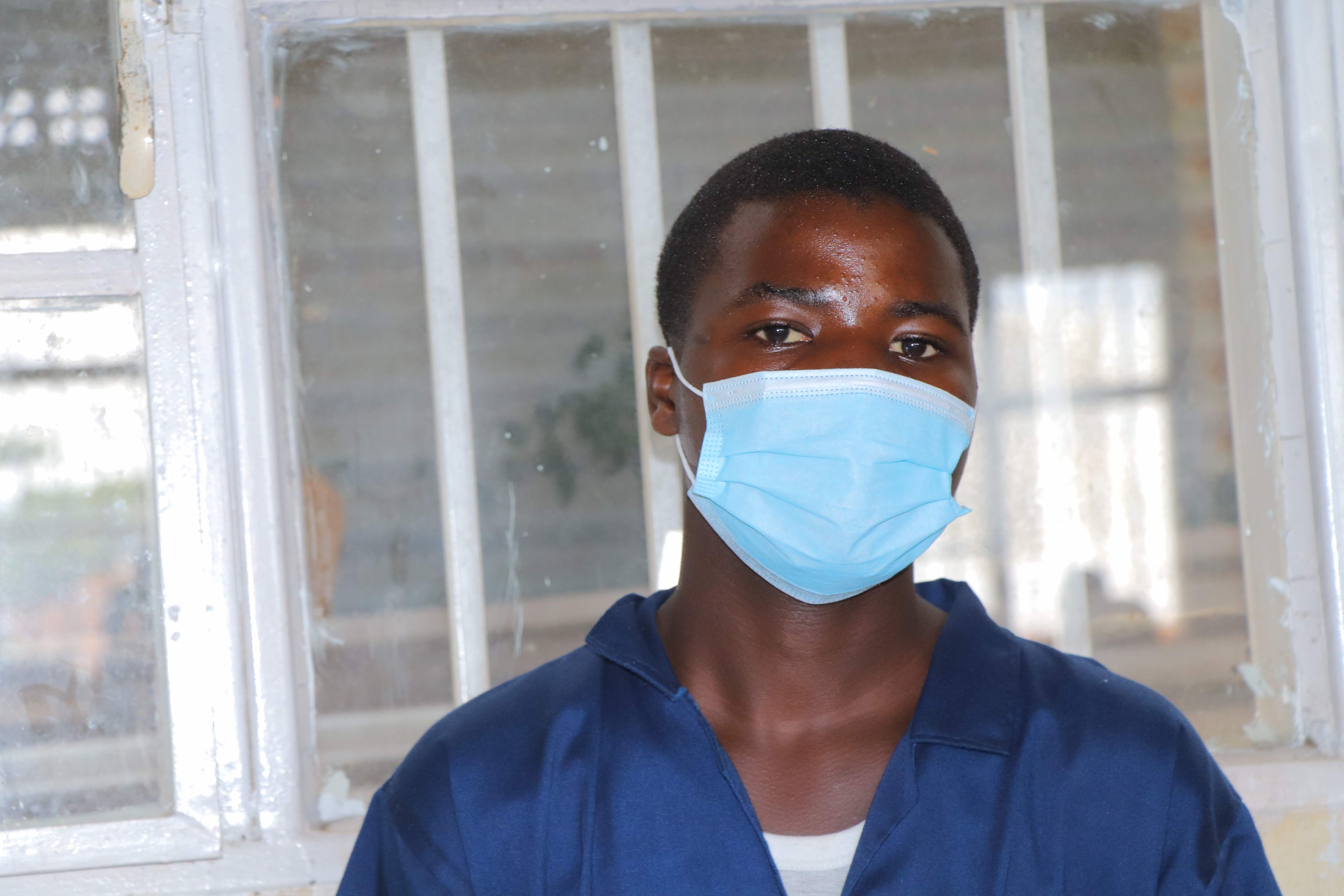 A person wearing a surgical mask and blue blouse looks into the camera, positioned in front of a large window
