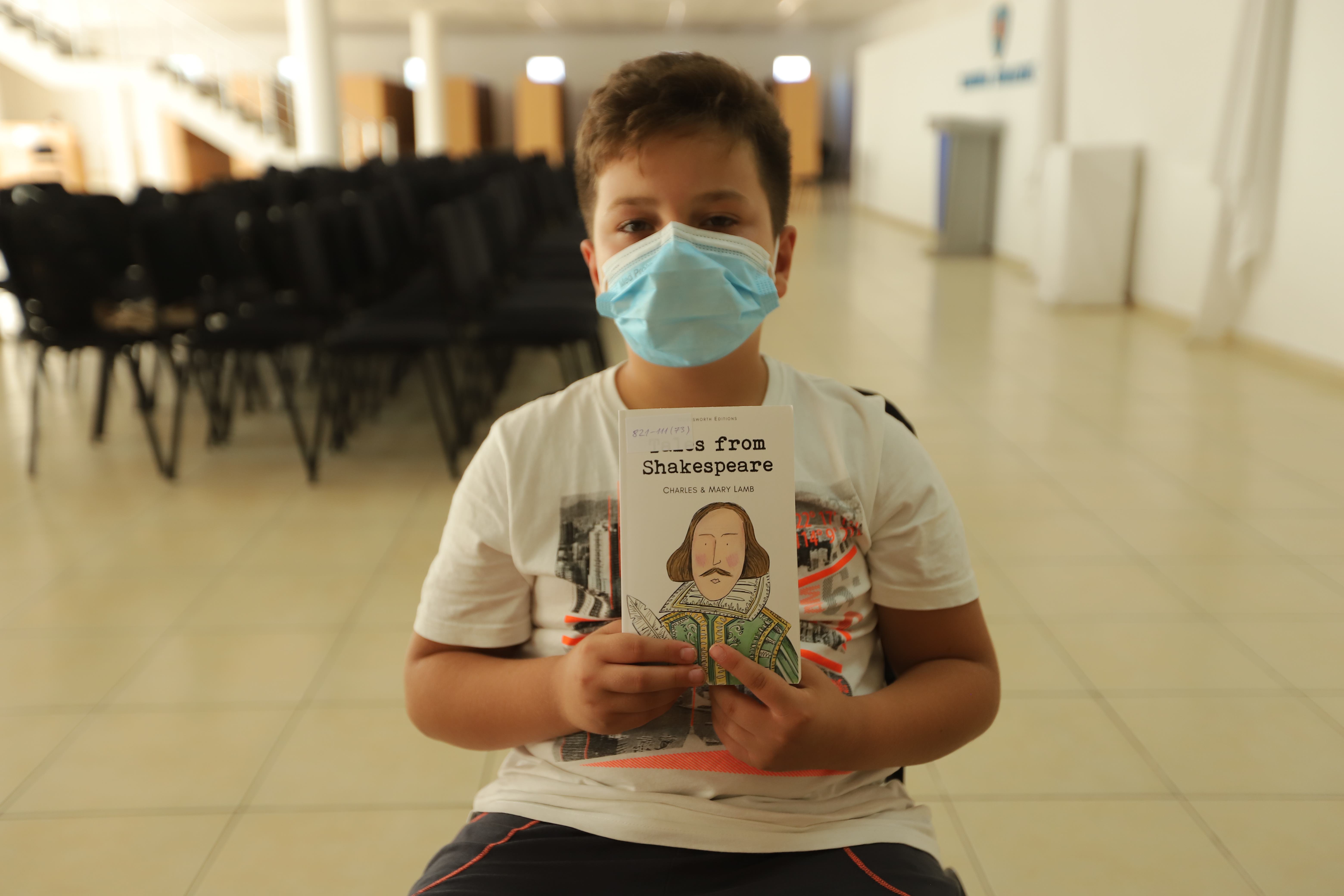 A young student in mask holds up a book with a hand drawn William Shakespeare on the cover