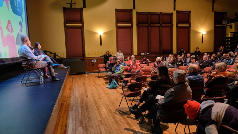 A large room filled with people looking at two people in chairs on a stage.