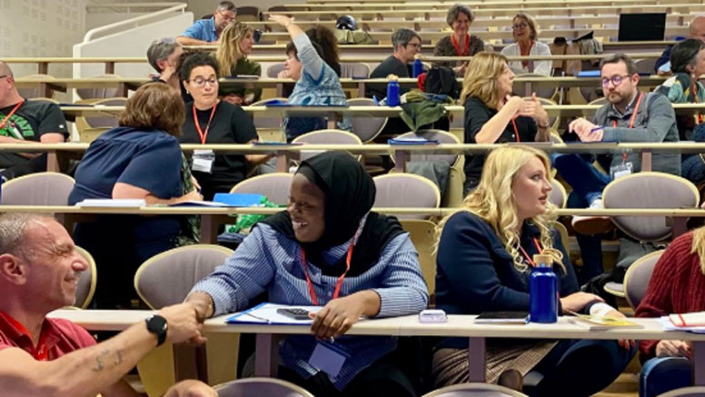 People in tiered desk seats at a conference in a classroom.