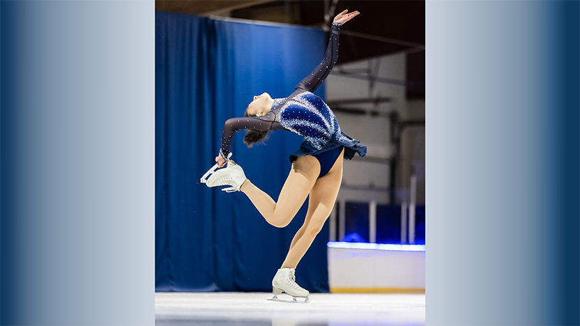 Ting Cui in blue and silver skating dress in the middle of a spin, bending backwards to grab one leg