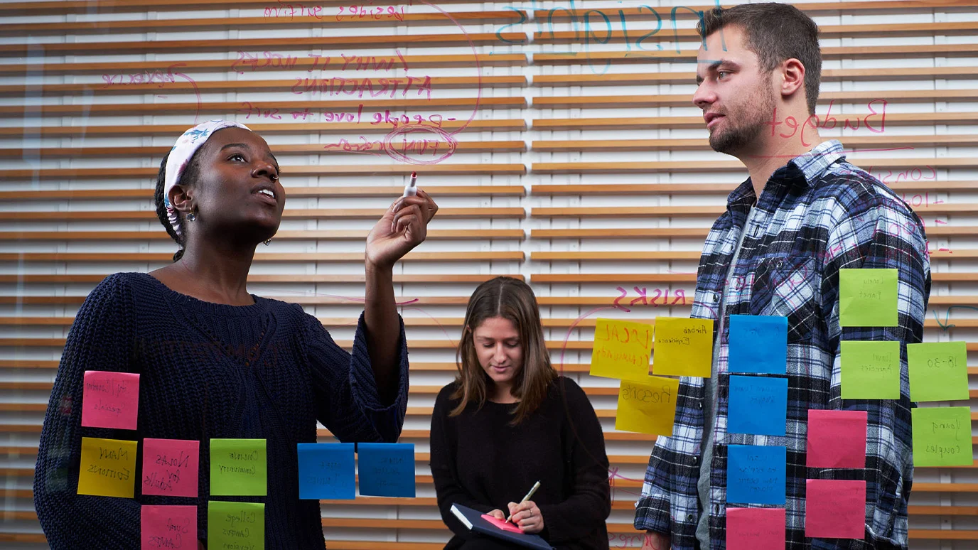 Three students using glass as a white board that also has many sticky notes on it.