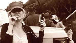 A woman stands in the foreground holding a piece of equipment. Behind her is a group of people with their backs to her looking at something else.