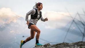 A woman is running on a mountain top with other large mountains in the background.