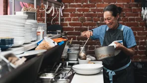 A woman in an apron is using a ladel to put soup in a bowl in a restaurant kitchen.