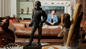 A woman sits infront of a firepace while looking at several small statues and figurines on a table in front of her.