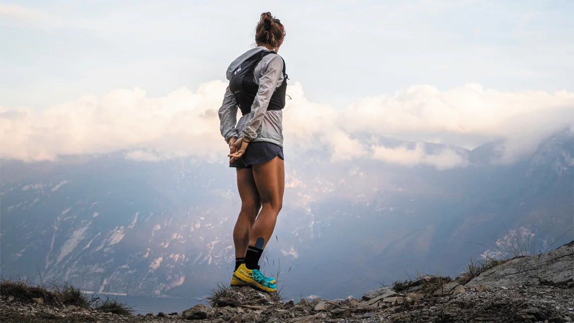 A woman stands on a mountain top looking at other mountains and a lake below.