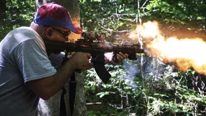 A man in a forest is firing an machine gun. Fire is coming out of the barrel.