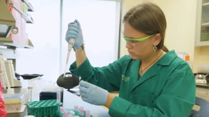 Clara Sandberg '24.5 is wearing a green lab coat and safety goggles and is working in a lab removing a sample from a test tube.