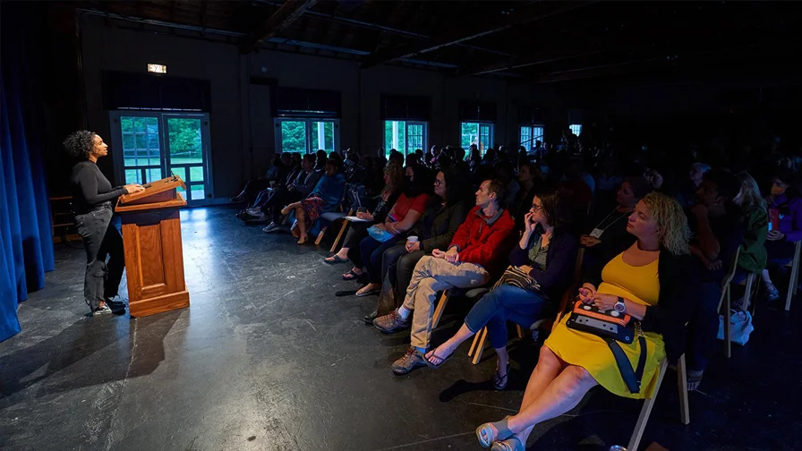 A woman speaks at a lectern infront of many attentive people sitting in a large room.