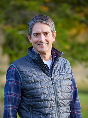 Tyler Newton outside at the Bread Loaf campus, in a dark plaid shirt and warm vest