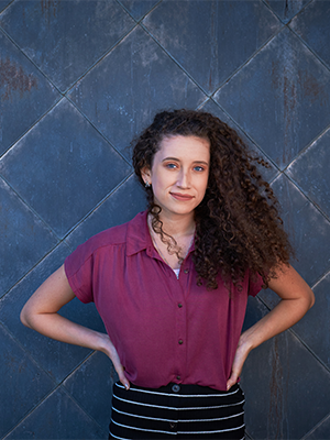 Zoe Garcia, wearing a short-sleeved plum button-up shirt, with hands on hips & loose shoulder-length curly hair