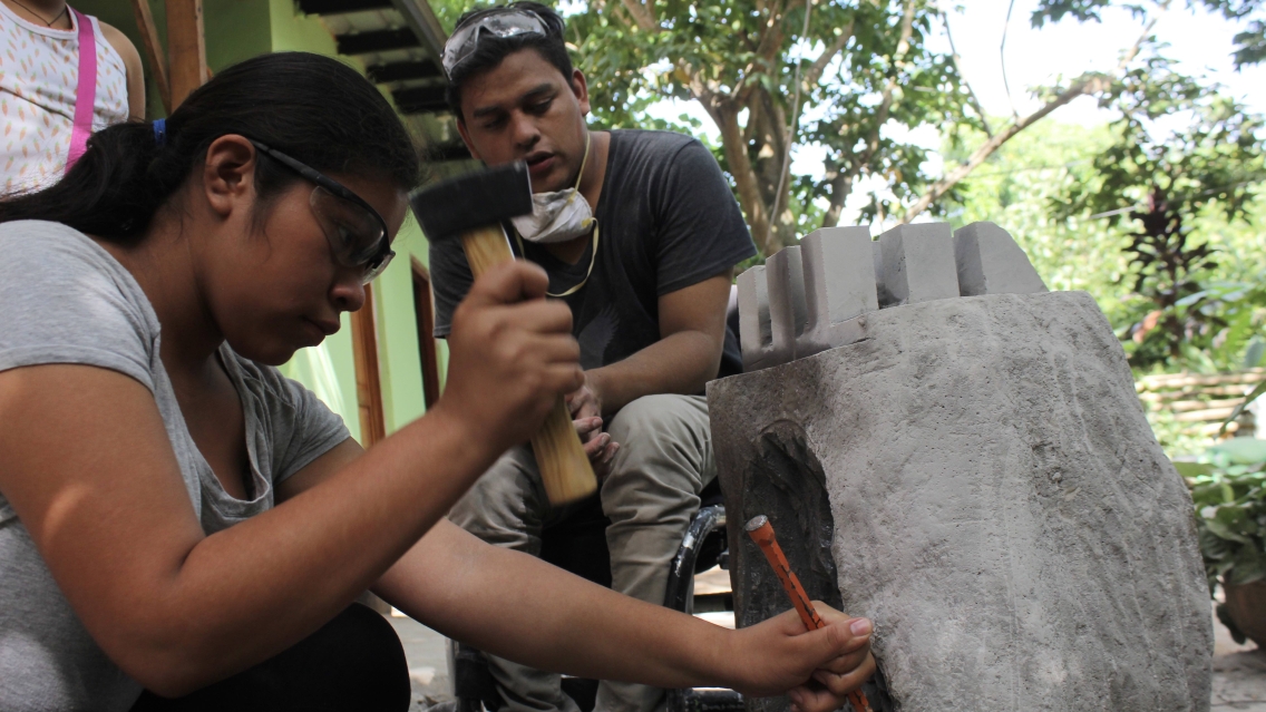 A man in a wheelchair supervises as a young girl sculpts a large rock