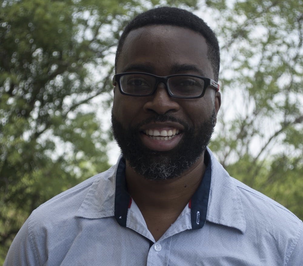 Neil Oculi smiles at the camera wearing a blue button-up and black glasses