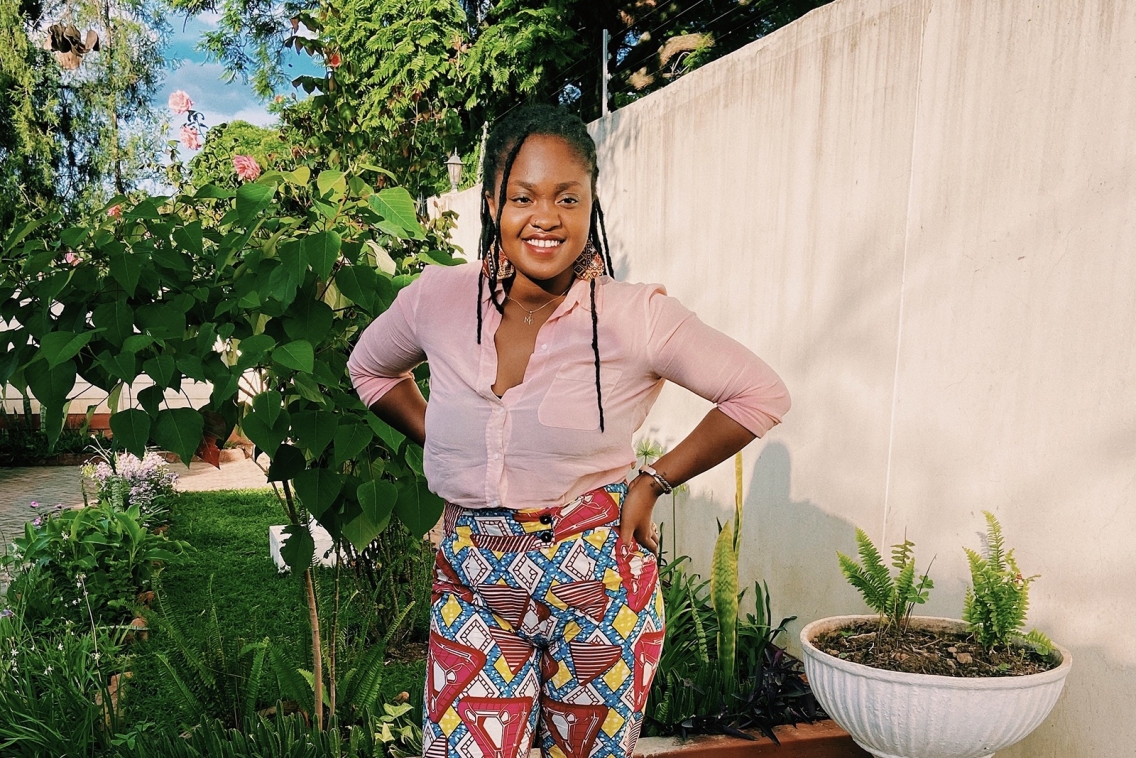 Marie-Simone Kadurira poses for the camera in a bright pink blouse and multi-colored pants in front of a tan fence and lush greenery.