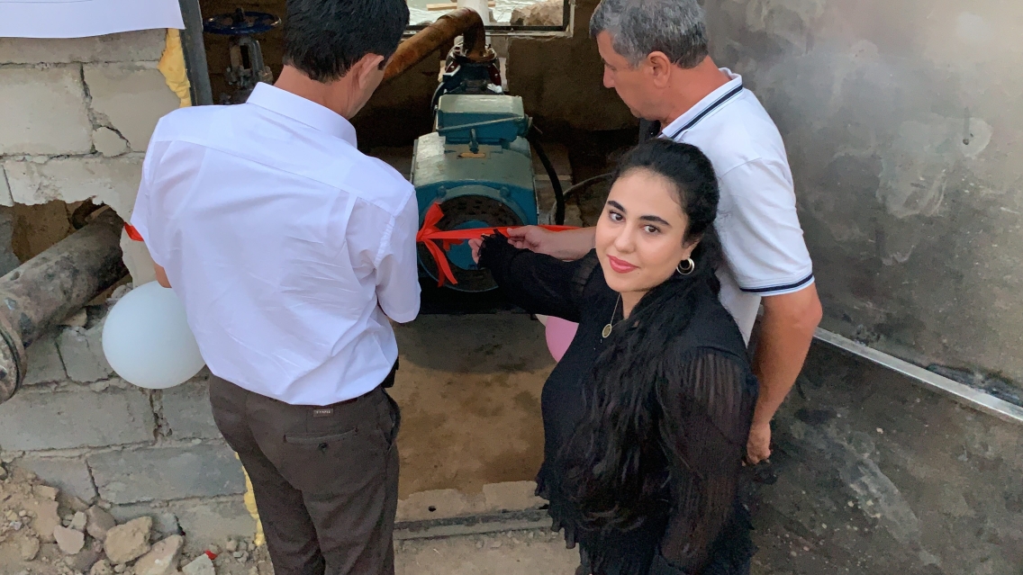 Amina Fatkhulloeva smiles at the camera flanked by two men working on a water pump in the midst of a concrete building