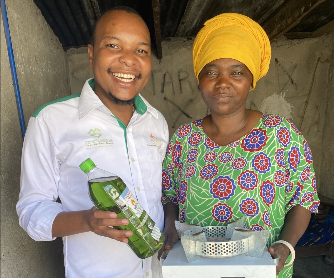 A man and woman pose together in front of a concrete wall. They hold a clean cookstove and a bottle of environmentally friendly cooking oil.