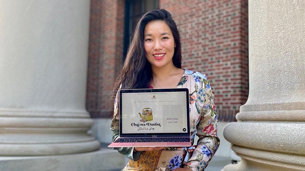 Lina Tori Jan sits and smiles next to a white pillar, holding an open laptop with a yellow image of a tea kettle and the words "Chai Wa Dastan" in black