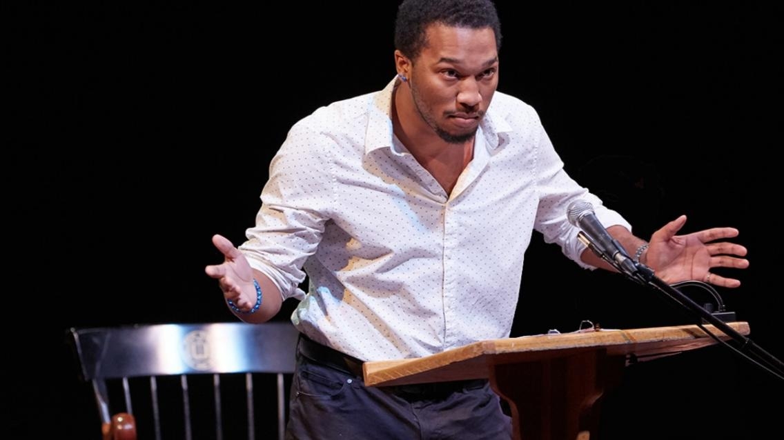 A college student in a white button-down and black pants gesticulates behind a podium while delivering a speech.