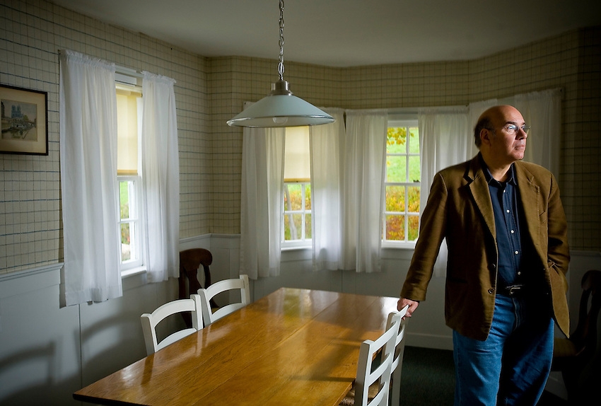 Jay Parini, wearing a brown suit jacket, blue shirt, and jeans, stands in a small dining room and leans on a light wooden table that has four white chairs tucked underneath it.