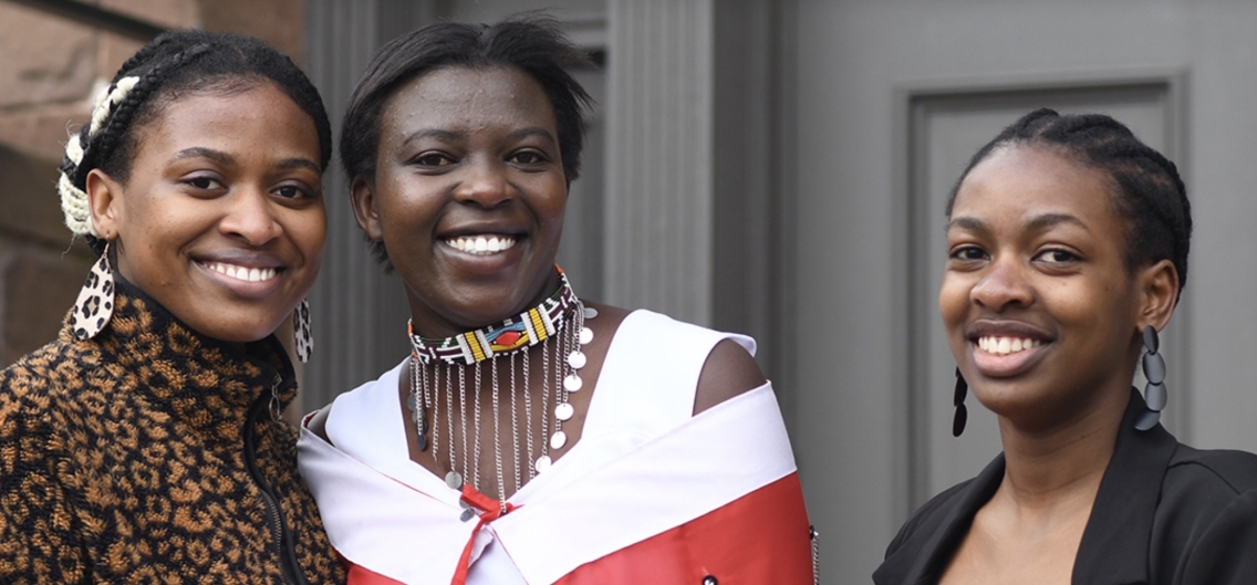 Constance Hirwa, Diana Naiyanoi Kimojino, and Alice Musabe pose together for a photo in front of a dark grey doorway.