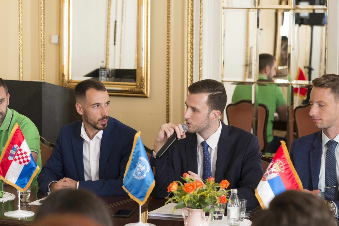 Wearing a dark suit and navy tie, Stanislav Knezevic sits behind a dark wooden desk adorned with country flags and speaks into a microphone.