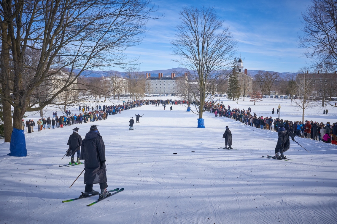 Six skiers in black graduation caps and gowns zip down a snowy hill surrounded by a ring of parents and families cheering them on.