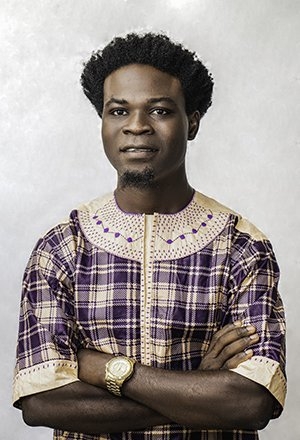Joseph Kaifala, wearing a cream and purple shirt, crosses his arms and poses in front of a white background.