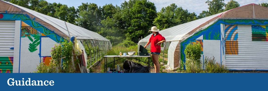 A person wearing tan shorts, a red sweater, and a tan bucket hat cleans paintbrushes at a table between two multi-colored, warehouse-style greenhouses. Below the picture a blue graphic footer features the word "Guidance" in white.