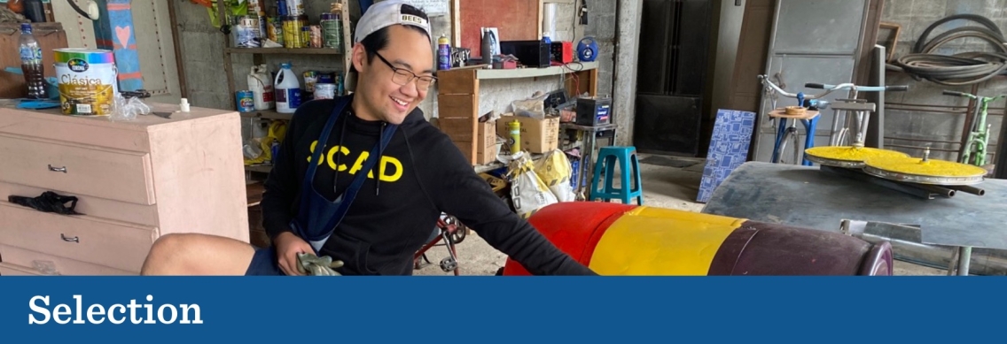 A young man wearing a black sweater with the word "SCAD" in yellow print smiles and leans over to touch a multi-colored painted barrel in the center of a cluttered mechanic workshop. Below the picture a blue graphic footer features the word "Selection" in white.