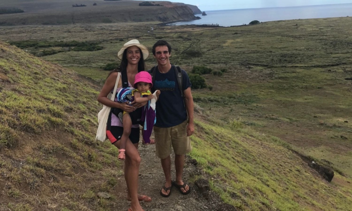 A woman wearing shoets, a tank top, her baby in a carrier, a white hat, and a white bag stand on a trail next to a man with a blue t-shirt and khaki shorts. They are both wearing flip flops, smiling into the camera, and a beautiful natural vista is behind them. You can see a green and brown landscape with a view of the ocean.