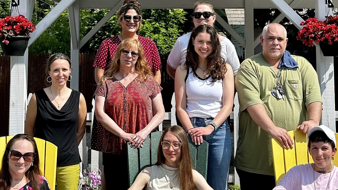 A group of nine people in three rows smile for the camera, three sit in Adirondack chairs. Sunny day in summer. 