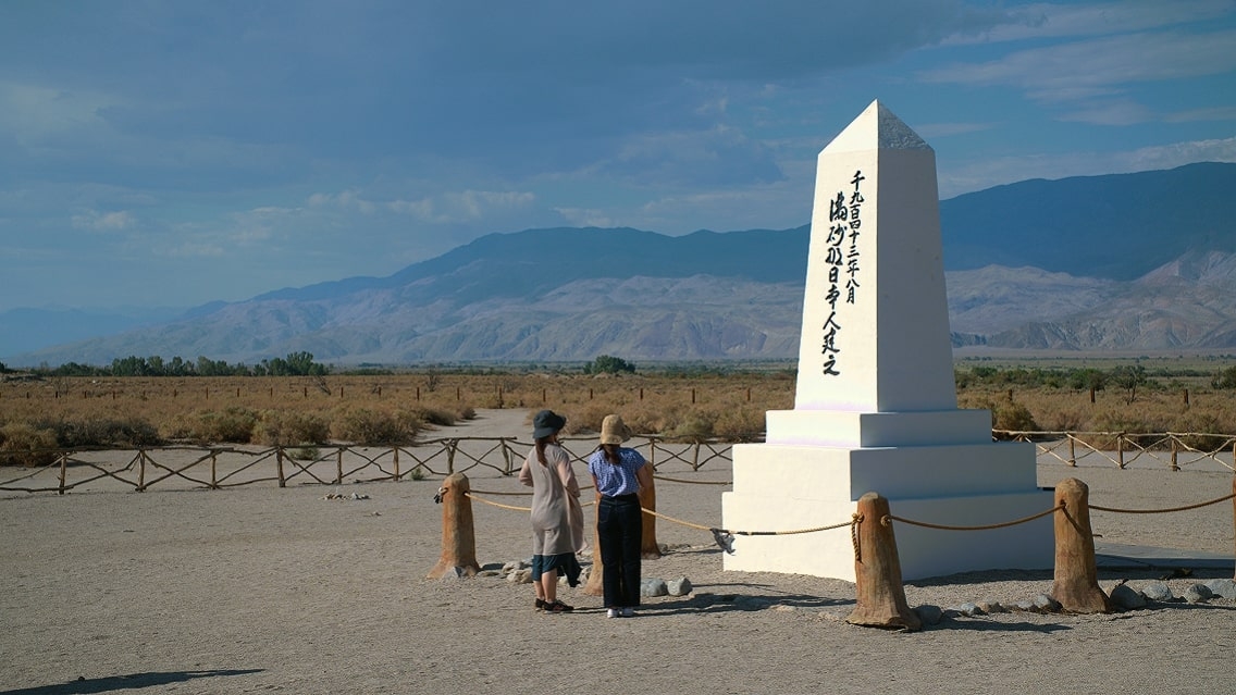 Two people stand in front of a white monument with Japanese lettering. The monument has a rope in front, and there is a blue sky and blue mountains in the background.