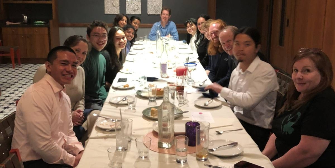 A group of students sitting at a dinner table. They are all turning to the camera and smiling. The table is set.